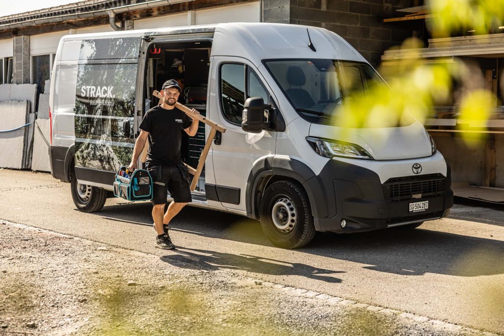 Handwerker mit Holzlatte vor dem beschrifteten Kastenwagen