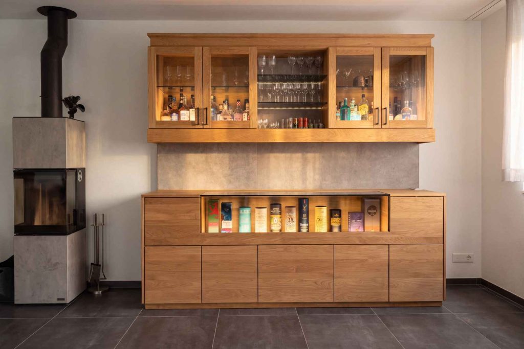 Whiskybar aus Eiche massiv im Wohnzimmer neben dem Cheminée, Vitrine und Hängeschränke beleuchtet