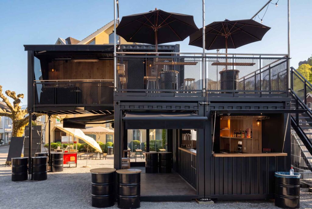 Zweigeschossige Container-Bar mit aufgeklappter Front, Dachterrasse und Fassstehtischen