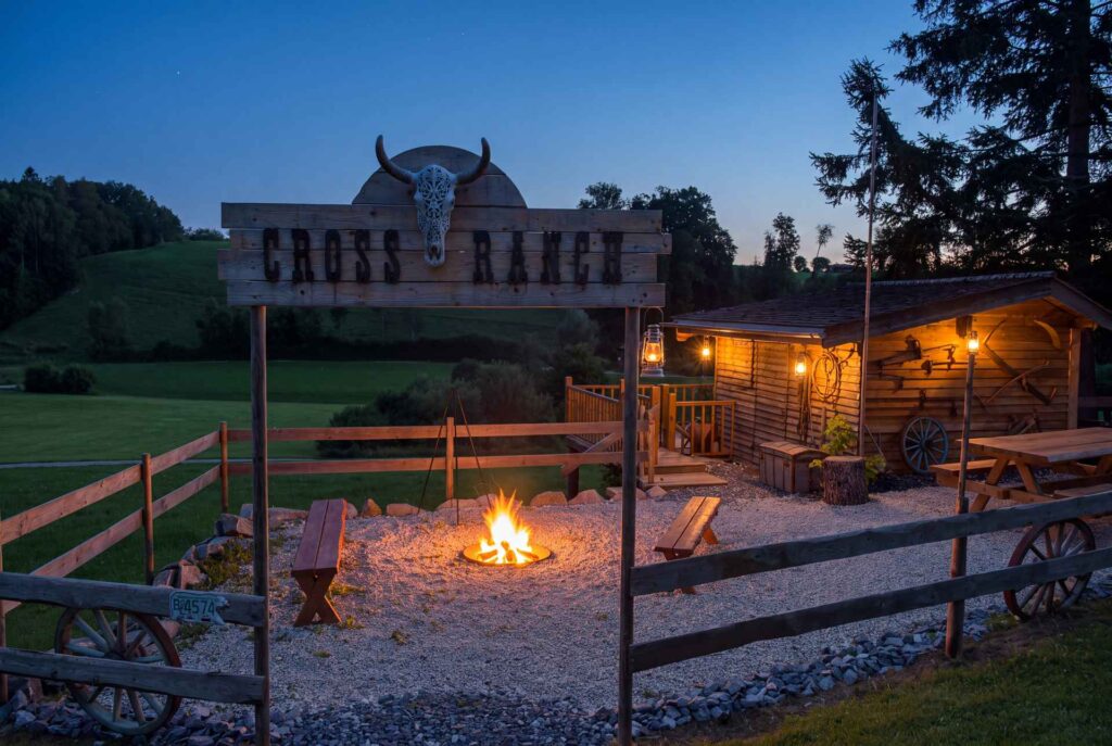 Themenbau im Westernstil von STRACK Baumanufaktur mit Veranda und brennender Feuerstelle in der Abenddämmerung