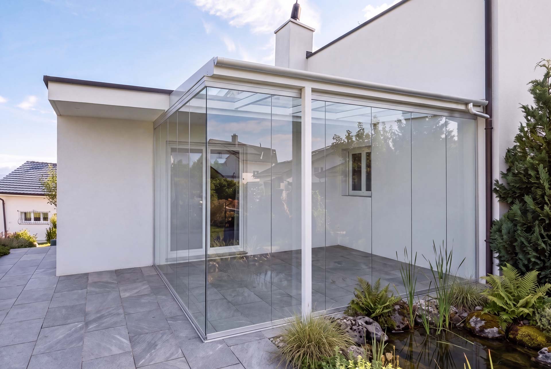 Rahmenlose Glasschiebewand mit Glasdach an der Hausecke, Natursteinplatten und Gartenteich davor